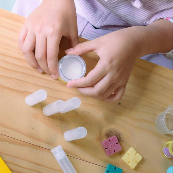 A person engaged in a DIY project with small containers and colorful blocks on a wooden table is opening a round container from the KISS NATURALS DIY Natural Lip Balm Kit, surrounded by several white cylindrical tubes and various non-toxic ingredients.