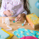 A child is engrossed in a DIY project, creating homemade lip balms at a wooden table. Utilizing the KISS NATURALS DIY Natural Lip Balm Kit, which includes small bottles and colorful containers, they follow instructions from a yellow box surrounded by scattered materials, all set against a pastel green wall.