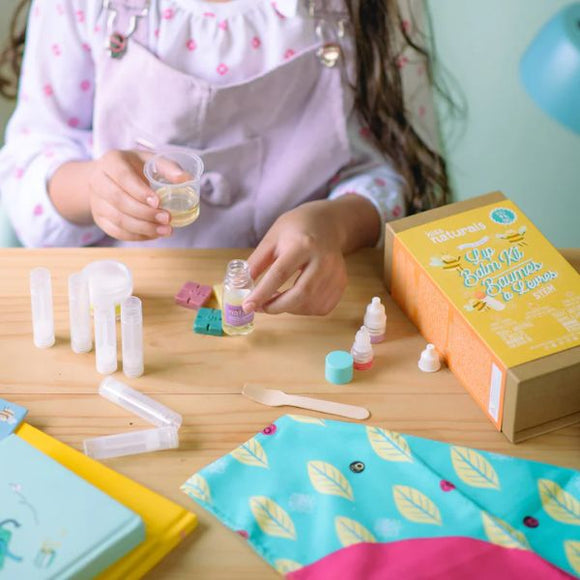 A child is engrossed in a DIY project, creating homemade lip balms at a wooden table. Utilizing the KISS NATURALS DIY Natural Lip Balm Kit, which includes small bottles and colorful containers, they follow instructions from a yellow box surrounded by scattered materials, all set against a pastel green wall.