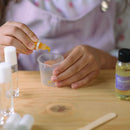 A person wearing a light purple apron uses a dropper to add liquid into a small plastic measuring cup. Bottles of oil and other containers are on the wooden table. The scene suggests a DIY activity involving all-natural ingredients or homemade products, possibly even crafting cruelty-free lip gloss using the DIY Natural Lava Lip Gloss Kit by KISS NATURALS.