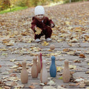 A child in a white hat and maroon outfit plays with the Korko Little Skittles Bowling Set outdoors, rolling a ball towards six colorful pins on a path of autumn leaves, enhancing their hand-eye coordination.