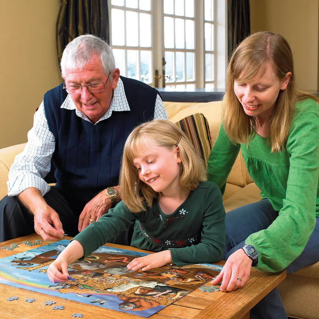 A smiling family collaborates on a Cobble Hill "Voyage of the Ark" jigsaw (350 pieces) while sitting on a beige sofa. An elderly man in a sweater vest, a young girl, and a woman in a green shirt fit pieces together. Bright light streams through large windows with white curtains behind them.