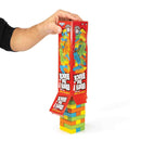 A hand holds a box of "Obey The Block" by Outset Media above a wobbly tower made of multicolored solid wood blocks, all set against a white background.