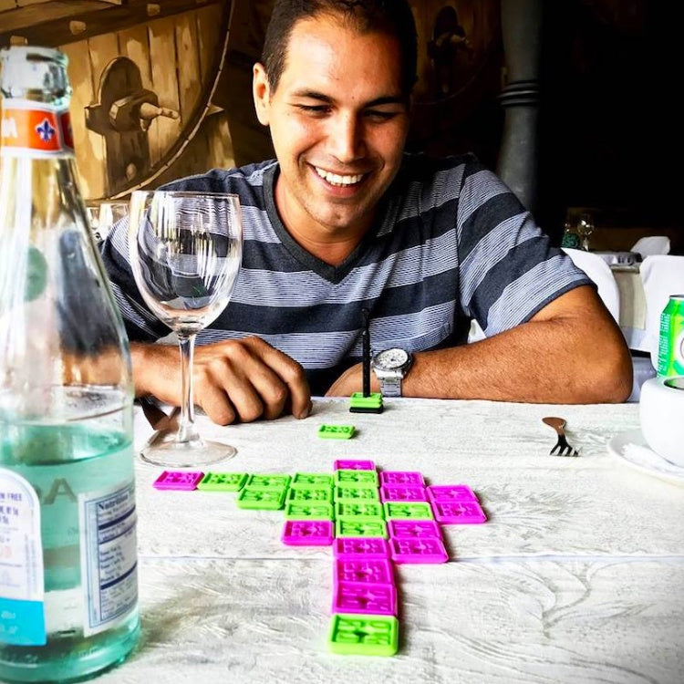Man sitting with a wine glass in front of him playing the OK Play game using green and pink tiles.