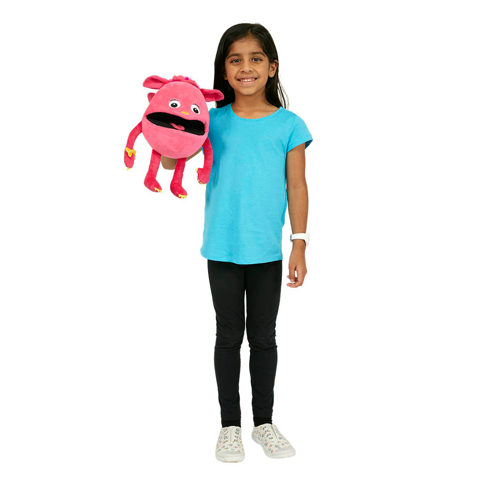 A young girl in a blue shirt and black pants smiles, holding THE PUPPET COMPANY's large Pink Baby Monster Puppet with eyes and a mouth, against a plain white background. Her excitement is as vibrant as the squeaky voice you'd imagine it having.