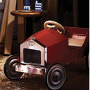 The Vilac Large Pedal Car in red, featuring a vintage style with a chrome grille and black wheels, rests on a dark floor sprinkled with wood shavings. A round wooden table with small buildings sits nearby, illuminated by ambient lighting that sets the scene for young drivers' classic adventures.