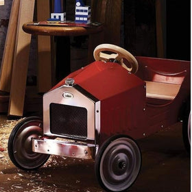 The Vilac Large Pedal Car in red, featuring a vintage style with a chrome grille and black wheels, rests on a dark floor sprinkled with wood shavings. A round wooden table with small buildings sits nearby, illuminated by ambient lighting that sets the scene for young drivers' classic adventures.