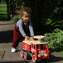 A toddler joyfully pushes a sustainable Vilac Ride-On Fire-truck outdoors on a sunny day, wearing red overalls and a striped shirt, with grass and plants in the background.