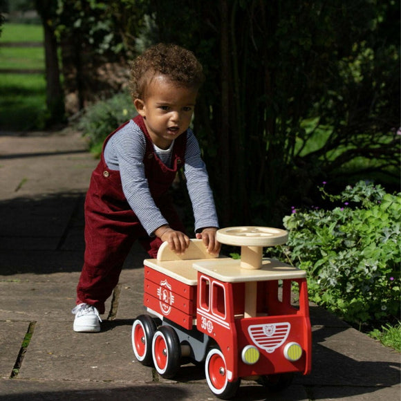 A toddler joyfully pushes a sustainable Vilac Ride-On Fire-truck outdoors on a sunny day, wearing red overalls and a striped shirt, with grass and plants in the background.