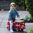 Outdoors on a stone patio with lush plants and steps in the background, a toddler gleefully rides the Vilac Ride-On Fire-truck, dressed in a blue cardigan and white tights.