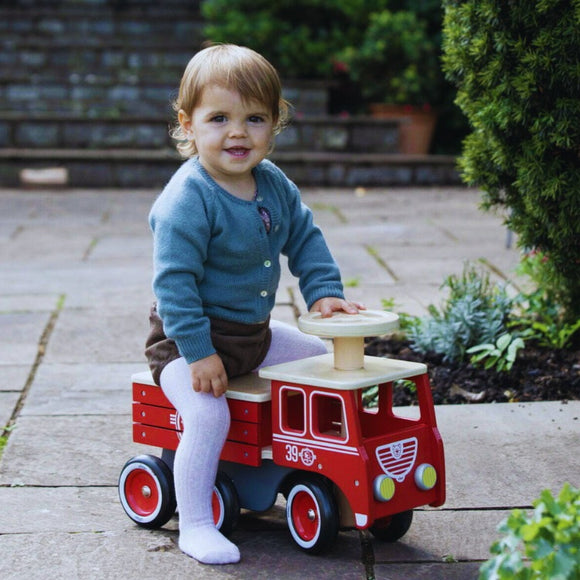 Outdoors on a stone patio with lush plants and steps in the background, a toddler gleefully rides the Vilac Ride-On Fire-truck, dressed in a blue cardigan and white tights.