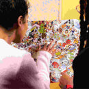 Two people are examining the Riddle Pics - Mayhem in the Library poster from Big Potato, which is filled with a vibrant array of cartoon characters and objects. The scene is bustling with activity, akin to an intricate search through visual riddles, suggesting an engaging exploration experience.