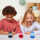 Excitedly, two children watch three Sensory Motion's Sensory Egg Volcano toys—red, black, and blue—on a table with swirling patterns inside for stress relief. Shelves and a round wall clock decorate the room's background.