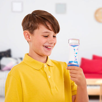 A young boy in a yellow polo shirt smiles as he holds a Sensory Motion Bubbler, showcasing its mesmerizing blue droplets and embracing mindfulness. The softly blurred room in the background, adorned with cushions and wall decorations, creates a serene atmosphere ideal for stress relief.