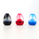 Three Sensory Motion's Sensory Egg Volcanos rest on a white surface. These egg-shaped fidget toys have a transparent speckled top and a solid-colored bottom: black, red, and blue from left to right. Ideal for stress relief.