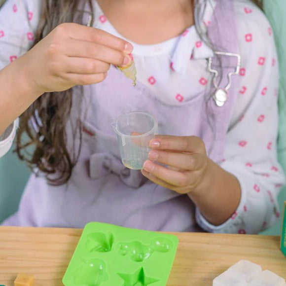 A child is using a dropper to pour liquid into a small cup, part of their DIY Natural Glycerin Soap Kit from KISS NATURALS. They are wearing a white shirt with red patterns and a light purple apron. On the table in front of them, there is a green silicone mold tray with various shapes for their DIY soap-making adventure.
