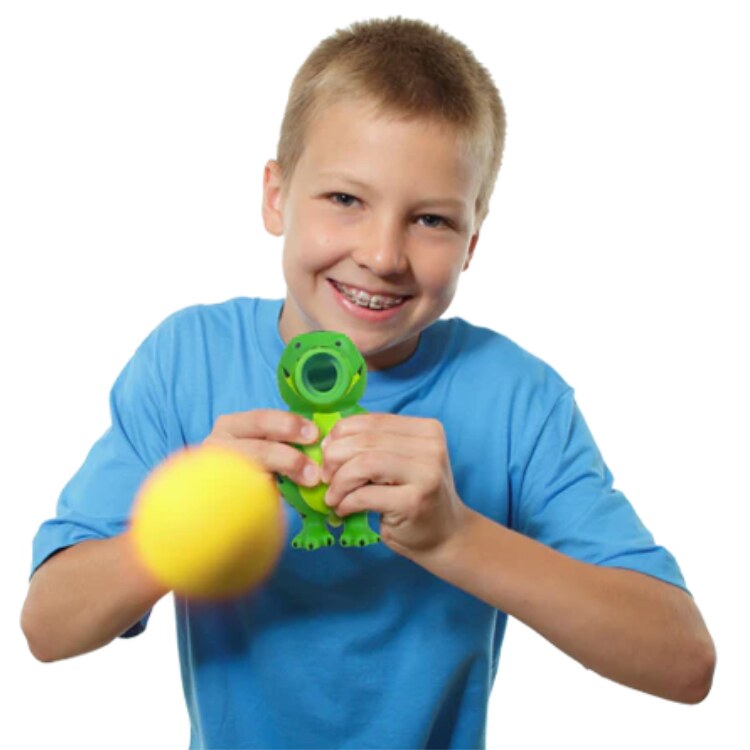 A young boy in a blue shirt smiles while holding a green dinosaur toy. He playfully launches a yellow foam ball from the Cheatwell Games Squeeze Popper: Dino towards the camera, making it perfect for indoor and outdoor play.