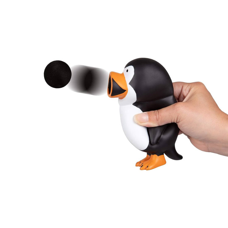 A hand squeezes the Cheatwell Games Squeeze Popper: Penguin, propelling a black foam ball from its beak. Ideal for both indoor and outdoor use, this penguin features a white belly, black back, and orange feet. The ball is seen midair against a plain white background.