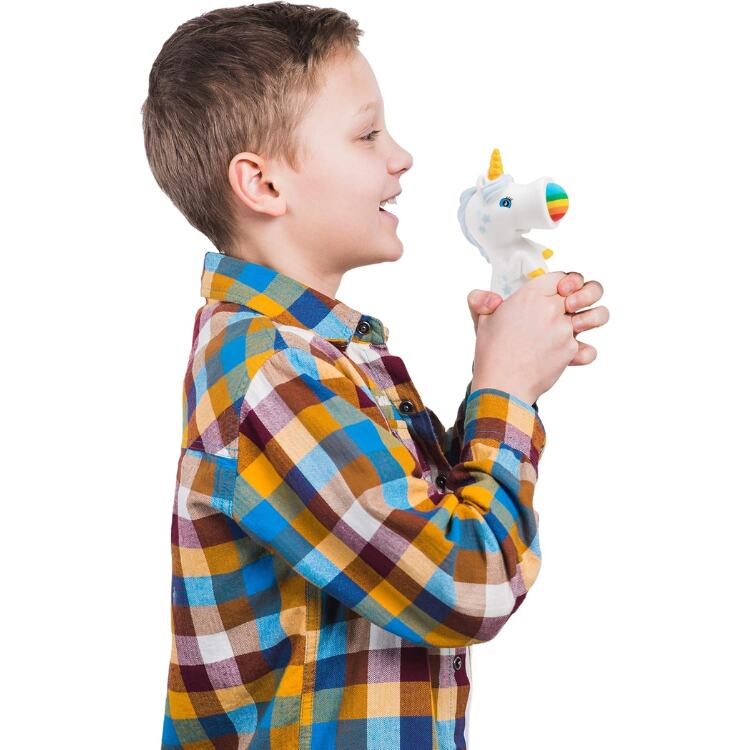 A smiling child in a vibrant plaid shirt joyfully squeezes the Cheatwell Games Squeeze Popper: Unicorn White, launching foam balls into the air as it lights up with rainbow colors.