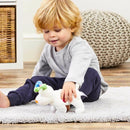 A young child with blonde hair sits on a soft white rug playing with "The Snowdog," a cuddly bean toy from Rainbow Designs, inspired by "Snowman and the Snowdog." The child wears a light gray long-sleeve shirt and navy pants, while a woven basket and gray knitted cushion sit in the background.