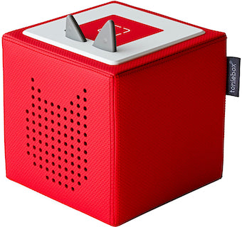 The Toniebox - Starter Set Red by Tonies is a red, cube-shaped audio system designed for children. It has two pointy, gray protrusions on top that resemble antennae and a perforated pattern on the side that looks like ears. A fabric tag labeled "tonies" is attached to the corner, making it ideal for sparking young imaginations during play.