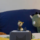 A small Tonie figurine of a cat with a star on its face stands on a gray, box-like Tonies speaker, poised to play the "Favourite Children's Songs - Bedtime Songs & Lullabies." Behind it, a green plush dinosaur leans against a blue pillow, ideal companions for a soothing sleep routine. The background features faintly patterned bedsheets.