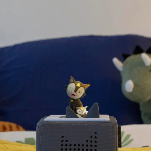 A small Tonie figurine of a cat with a star on its face stands on a gray, box-like Tonies speaker, poised to play the "Favourite Children's Songs - Bedtime Songs & Lullabies." Behind it, a green plush dinosaur leans against a blue pillow, ideal companions for a soothing sleep routine. The background features faintly patterned bedsheets.
