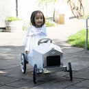 A young apprentice driver with shoulder-length hair smiles while sitting in a Vilac - Large Pedal Car - White. The outdoor setting, featuring stone pavement and lush greenery, adds to the timeless charm of this scene.