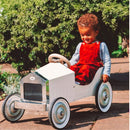 A young apprentice driver in red overalls joyfully pedals a Vilac - Large Pedal Car - White along a paved path surrounded by lush greenery, relishing its classic look.