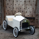 The Vilac Large Pedal Car in white is a vintage-style metal car featuring a nostalgic design, steering wheel, and chrome accents. It rests on stone before a rustic wooden door with metal details, poised to become a cherished family heirloom.