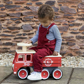 A toddler wearing a striped shirt and red overalls sits on the Vilac Ride-On Fire-truck. The child looks down with one hand on the steering wheel, against a backdrop of a stone wall and pebbles. This charming toy is crafted from sustainably-sourced wood by Vilac.