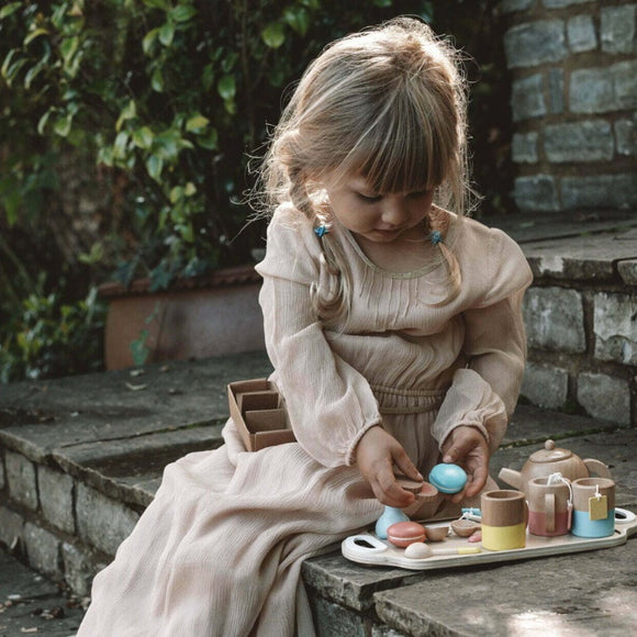 A young girl with braided hair sits on stone steps, enjoying her Vilac Wooden Tea Set. She wears a long, light-colored dress and is surrounded by greenery as she prepares wooden biscuits for her delightful children's tea party.