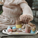 A child in a beige dress sits outdoors, cradling a wooden cup from the Vilac Wooden Tea Set. The set features pastel-colored cups, a teapot, and wooden biscuits resembling small pastries, all neatly arranged on a tray atop the stone surface.