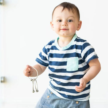 A smiling toddler with short dark hair is standing against a white background. The child is wearing a blue and white striped shirt with a light blue pocket, and blue striped pants. They are holding YUMMIKEYS' Yummirings - Stainless Steel Teething Rings, which resemble a small tuning fork.