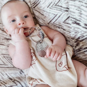 A baby with a curious expression plays with YUMMIKEYS' Stainless Steel Woodland Teething Keys in their mouth. Lying on a patterned blanket, they wear a cream outfit with red line drawings and are fully engaged with the sensory animal keys. The baby has light skin and short hair.
