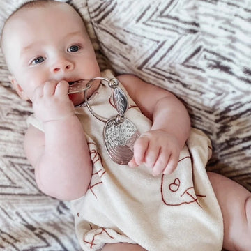 A baby with a curious expression plays with YUMMIKEYS' Stainless Steel Woodland Teething Keys in their mouth. Lying on a patterned blanket, they wear a cream outfit with red line drawings and are fully engaged with the sensory animal keys. The baby has light skin and short hair.