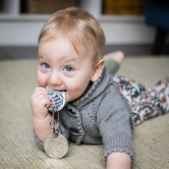A light-haired, blue-eyed baby lies on a soft surface, playfully holding YUMMIKEYS' Stainless Steel Woodland Teething Keys in their mouth. They wear a gray knit sweater and patterned pants, gazing upward with curiosity.