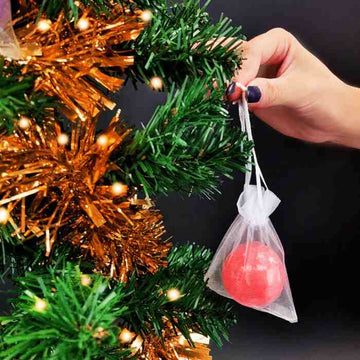 A hand gently places a Baff Bombz Bauble by Zimpli Kids onto the Christmas tree, adorned with orange tinsel and twinkling lights. The red biodegradable fabric sparkles, adding an eco-friendly touch to the festive display.