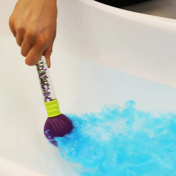 A hand stirs the blue water in a bathtub with Zimpli Kids' Baff Bombz Magic Brush, revealing its colorful, sparkly handle and purple bristles. The scene transforms into an enchanting, eco-friendly fizzing bathtime experience.