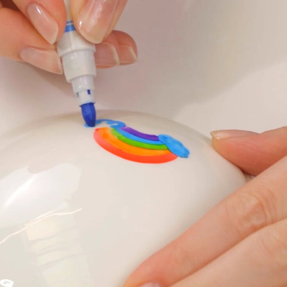 A close-up of hands drawing a small rainbow on a white surface using Zimpli Kids' Doodle 'N' Dip. The rainbow has red, orange, yellow, green, and purple bands with a blue cloud at each end.