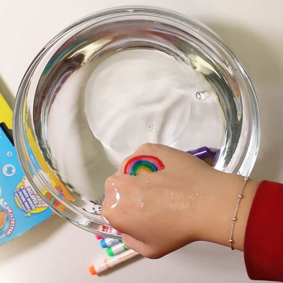A hand with a rainbow ring and unique tattoo dips into a clear bowl of water, surrounded by Zimpli Kids' Doodle 'N' Dip magic markers and a partially visible yellow object on the white surface.