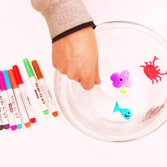 A hand holds a Zimpli Kids' Doodle 'N' Dip white spoon with a purple flower over a glass bowl of water displaying floating images of a red crab and blue whale. Nearby, magic markers await to enhance this vivid aquatic scene.