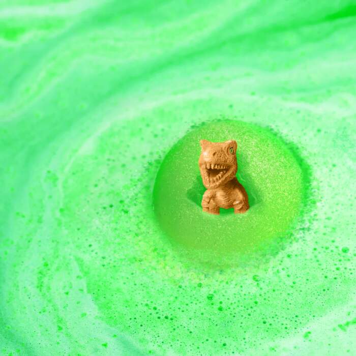 The Zimpli Dino Surprise Figure Baff Bombz by Zimpli Kids reveals a small, child-friendly toy dinosaur amid vibrant green foam, crafted by bubbling bath bombs for a whimsical and playful scene.