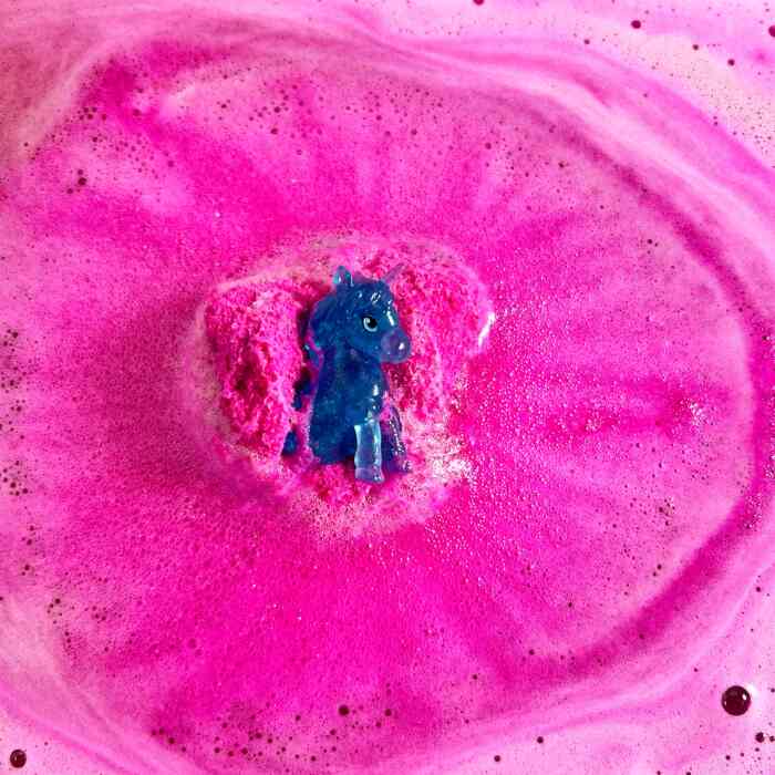 A Zimpli Unicorn Surprise Figure from Zimpli Kids is surrounded by vibrant pink foam and bubbling bath bombs in a swirling bath. The figure appears to be at the center, creating an eye-catching contrast with the bright pink background.