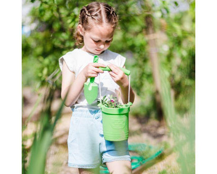 A young girl enjoys a sunny day in a lush garden with The Toy Shop's Eco Spade and green bucket made from wheat fiber. She's dressed in a white shirt and light blue shorts, ready to nurture her foldable activity toy buckets.