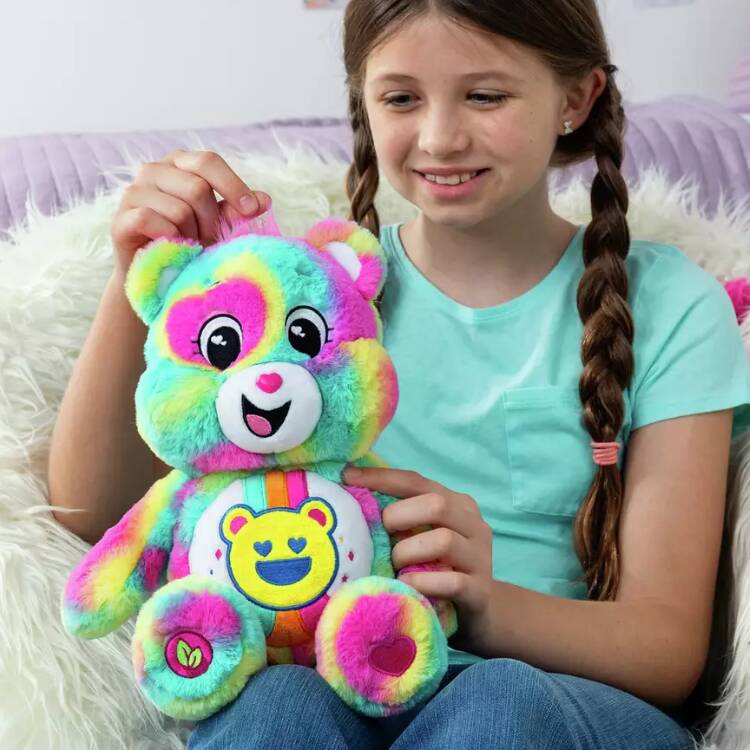 A girl with braided hair smiles while holding the 35cm Care Bears Eco-friendly Good Vibes Bear by Basic Fun, featuring heart and smiley face designs. She sits on a fluffy cushion in a cozy room.