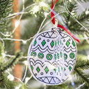 The Eggnogg Card Book - 3D Christmas Decorations by Eggnog features a handcrafted ornament with green and black geometric patterns hanging elegantly from a tree branch with white string lights. A festive red ribbon secures it in place, adding charm to this colorful holiday scene.