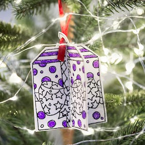 The Eggnogg Card Book from Eggnog features a hand-drawn 3D gift box ornament for coloring. Adorned with purple dots, stars, and a red ribbon, it hangs on the Christmas tree illuminated by white lights—perfect as a children's activity centerpiece.