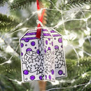 The Eggnogg Card Book from Eggnog features a hand-drawn 3D gift box ornament for coloring. Adorned with purple dots, stars, and a red ribbon, it hangs on the Christmas tree illuminated by white lights—perfect as a children's activity centerpiece.