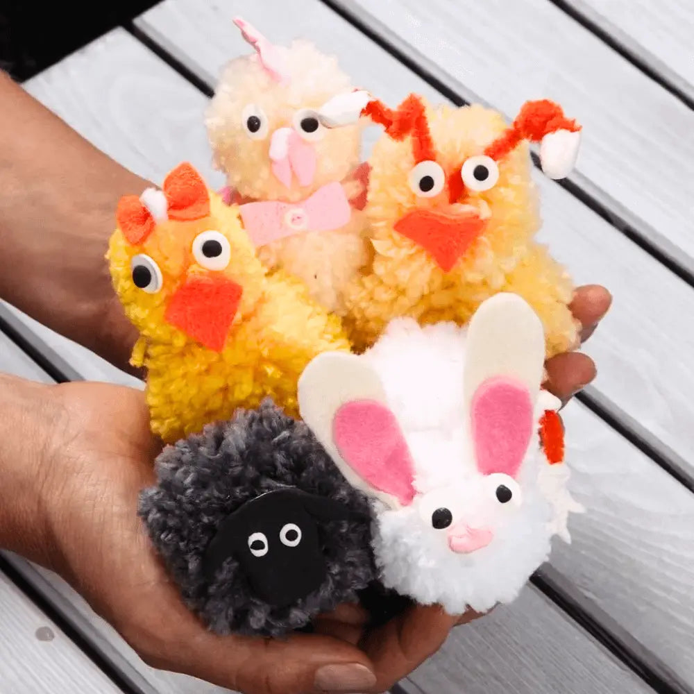 Crafted pom-pom animals - a black sheep, white bunny and 3 chicks held in two hands. Slatted wood behind.